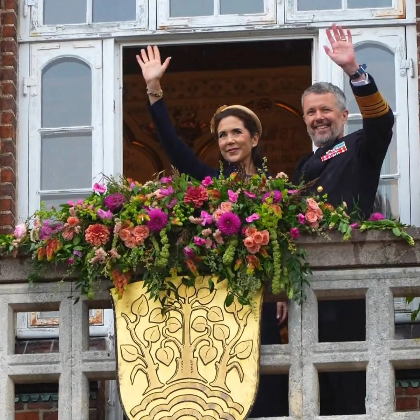 Kongen og dronningen hilser fra en balkon i Holbæk med Meretes farverige blomsterudsmykning foran sig